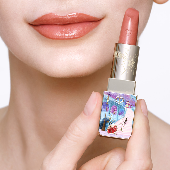 Photo of the 26 AW Holiday Collection limited-edition LIPSTICK on a fair-skinned model. A close-up of her lips is shown next to her hand holding an opened lipstick tube featuring a detailed scenic illustration with gold star details.
