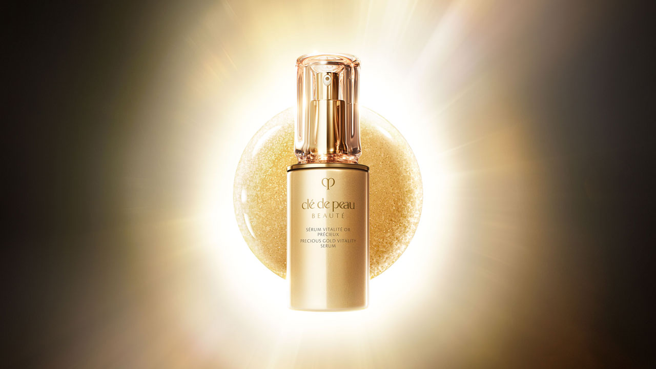 Hình ảnh tinh chất dưỡng da PRECIOUS GOLD VITALITY SERUM được chiếu sáng bởi ánh sáng vàng rực rỡ, đặt trên nền có họa tiết với các hạt vàng lấp lánh, tạo nên một hình ảnh sang trọng và đậm chất điện ảnh.