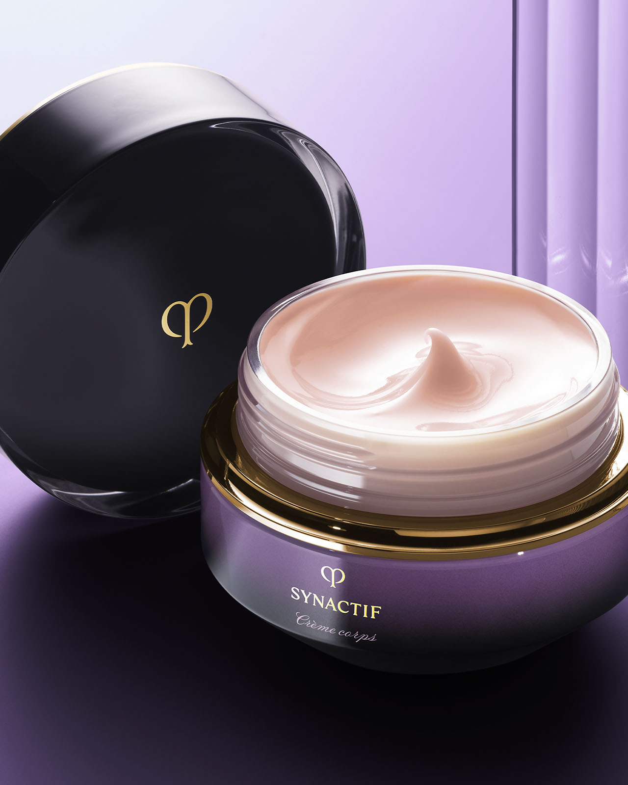 Photo of an open jar of SYNACTIF body cream, showing the soft, pale pink cream inside with its black cap leaning against it. The scene is set against a soft purple and white gradient background.